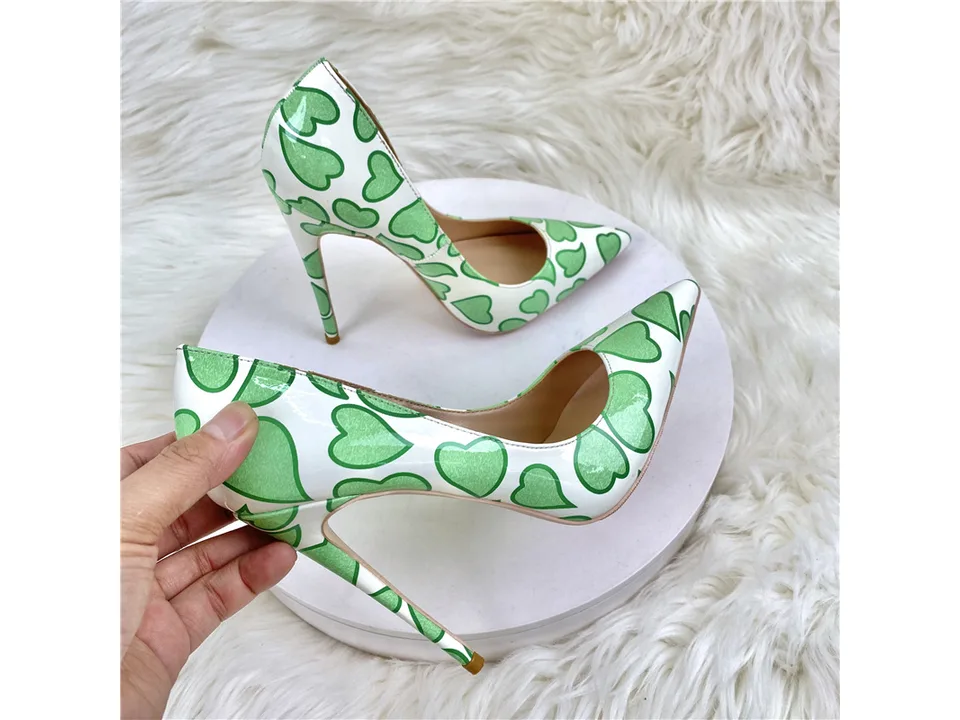 A hand holding one of the white and green heart-patterned stiletto pumps.