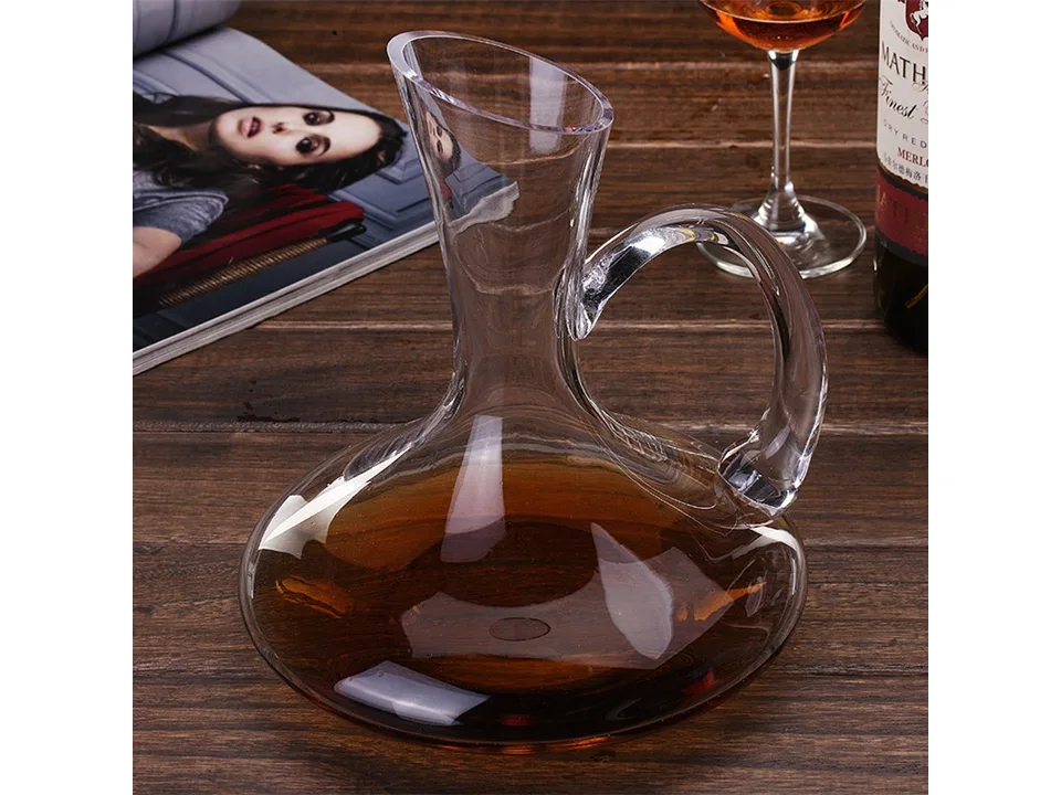 A glass wine decanter with a handle, filled with wine, sitting on a wooden table next to a wine glass.