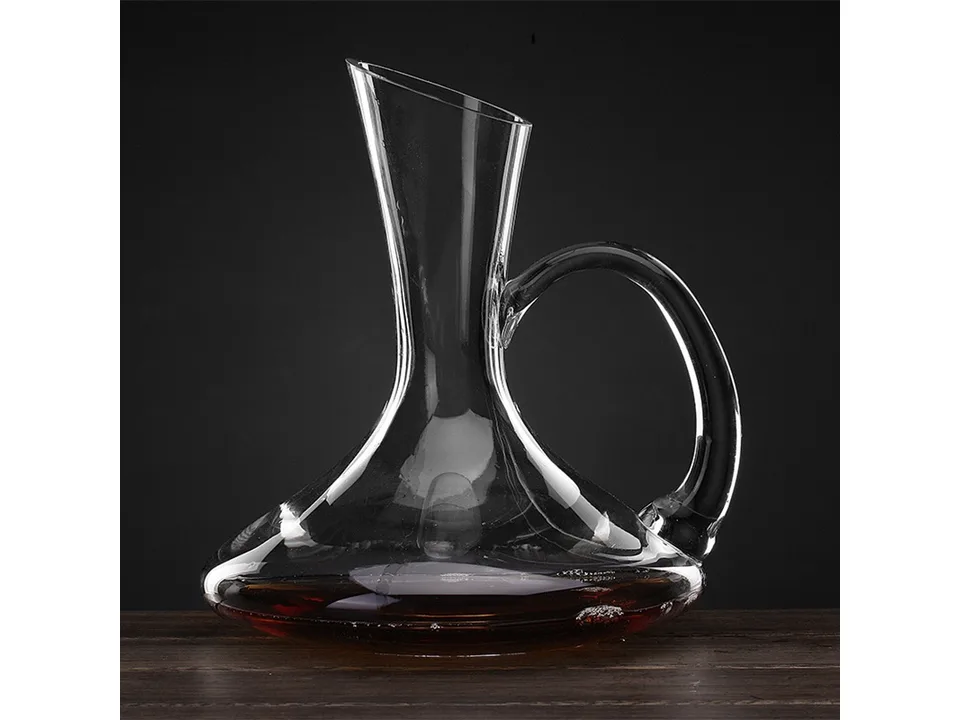 A clear glass wine decanter with a handle, filled with a small amount of red wine, on a wooden surface.