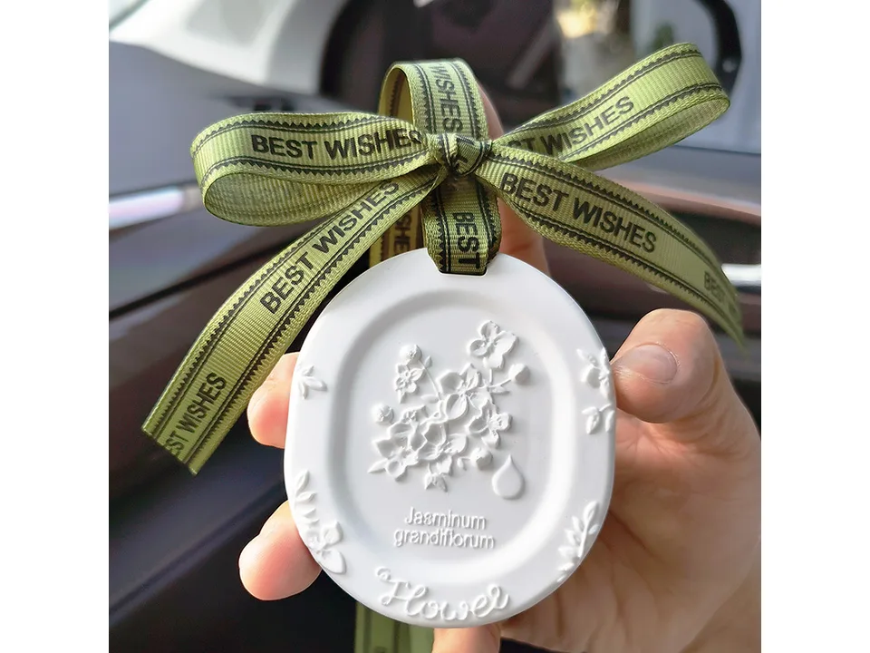 A hand holding a white, oval-shaped ceramic fragrance stone with a floral relief design and a green ribbon.