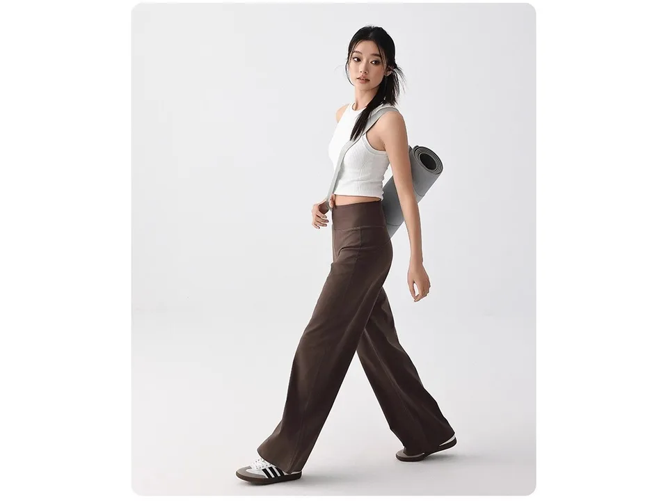 A model walking while wearing brown loose-fitting yoga pants and carrying a yoga mat.