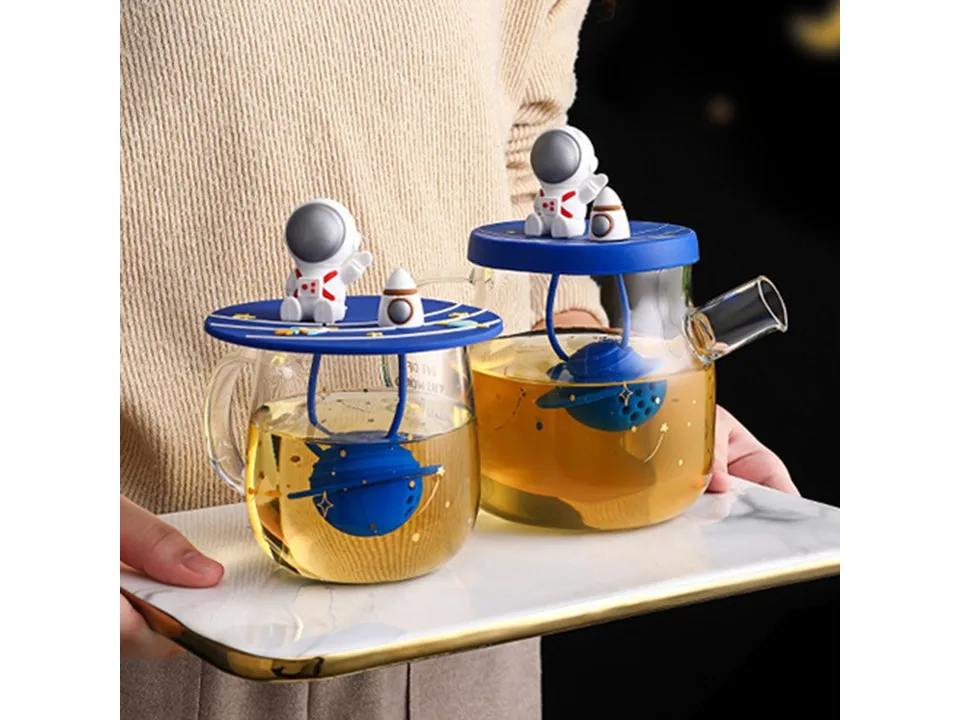 A person holding a glass teapot and a glass mug, both with astronaut-themed lids and tea infusers.