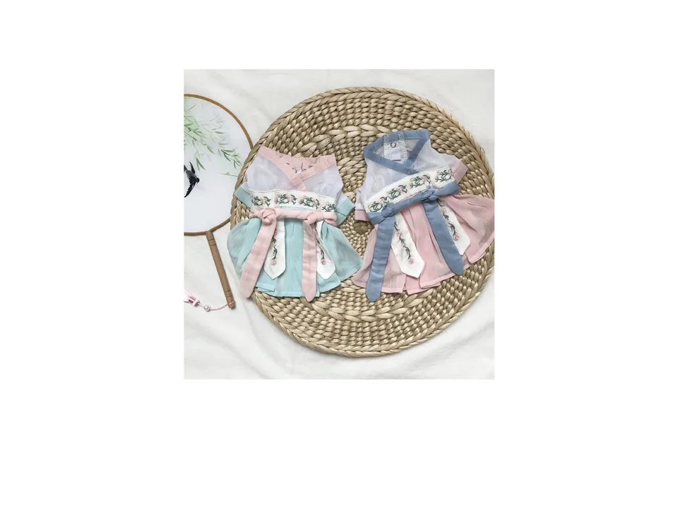 Two traditional style Hanfu dresses for pets, one in blue and one in pink, laid out on a woven mat.